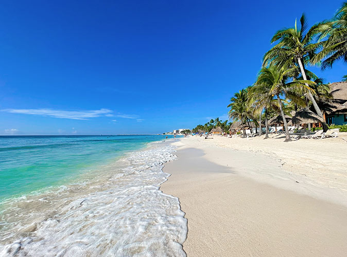 An idyllic looking white sandy beach, bright blue sea, some palm trees and some buildings in the distant background