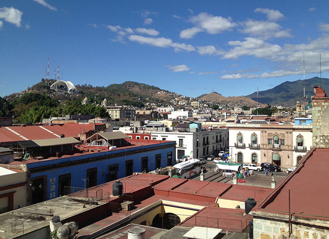 Life in Oaxaca An aerial view of Oaxaca, with many houses with red roofs, a town square and green hills in the background