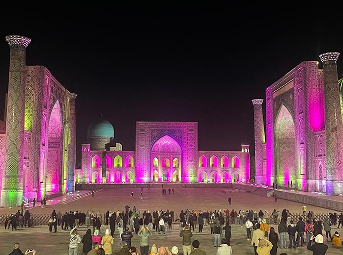 The Registan, a popular place to visit for people living in Uzbekistan A large historical brown building surrounding a courtyard with many people, lit up with pink lights