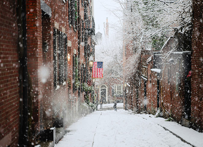 The cheapest time to visit Boston A snow covered street with brick buildings on both sides and an American flag