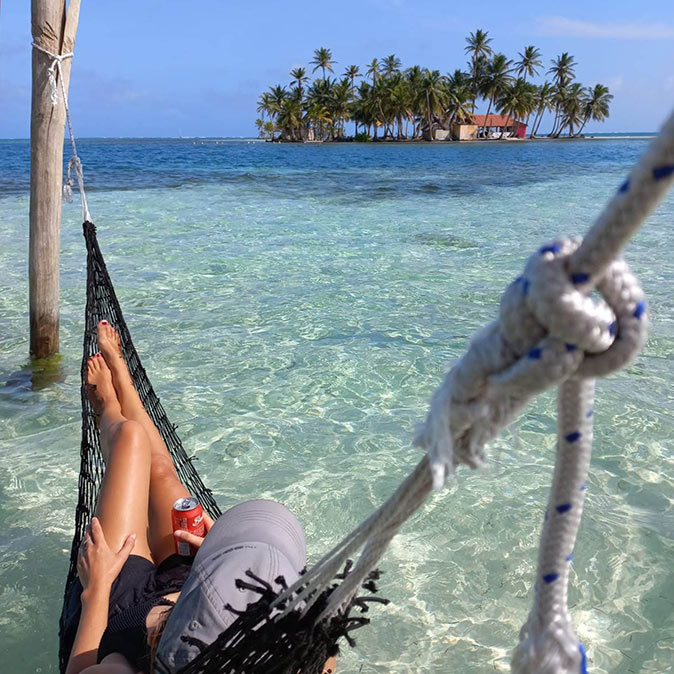 Traveling off the beaten path woman in a hammock holding a drink, with azure blue water and an idyllic tropical island in front of her