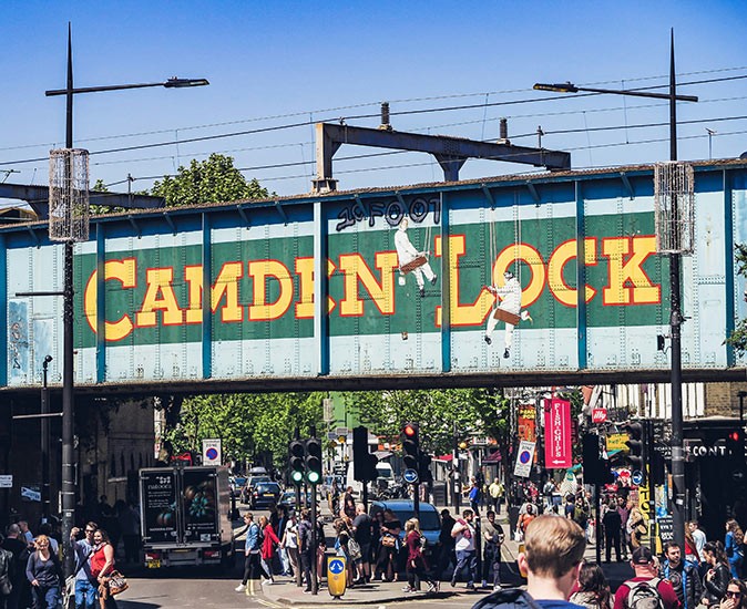 The famous Camden Lock sign, a popular site to see in Camden Town a street filled with pedestrians with a bridge over it with the famous Camden Lock sign
