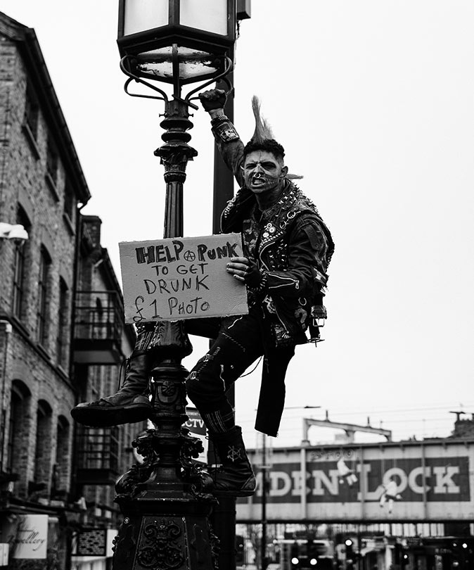 Visiting Camden Town, a unique part of London black and white picture of a man dressed in a punk outfit, climbed halfway up a street light, holding a a sign asking for money