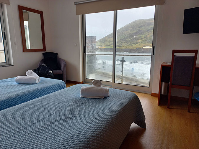 A bedroom with 2 single beds, a chair, a mirros and a large window with sea view