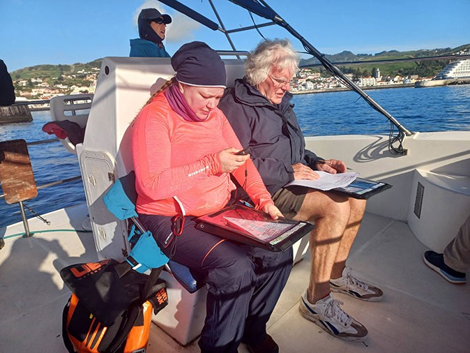 3 people on a boat, 1 writing something, 1 looking at a gps and 1 standing in the background