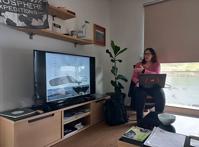 A woman with a laptop sitting next to a tv, giving a presentation about whales