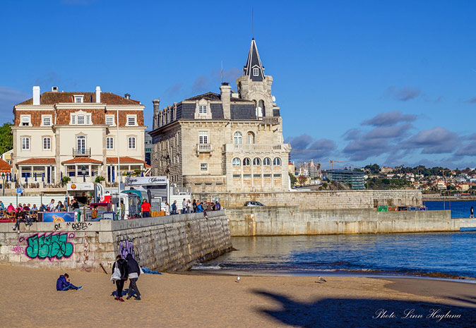 Several people on a beach with historical gray, brown and white buildings in the background