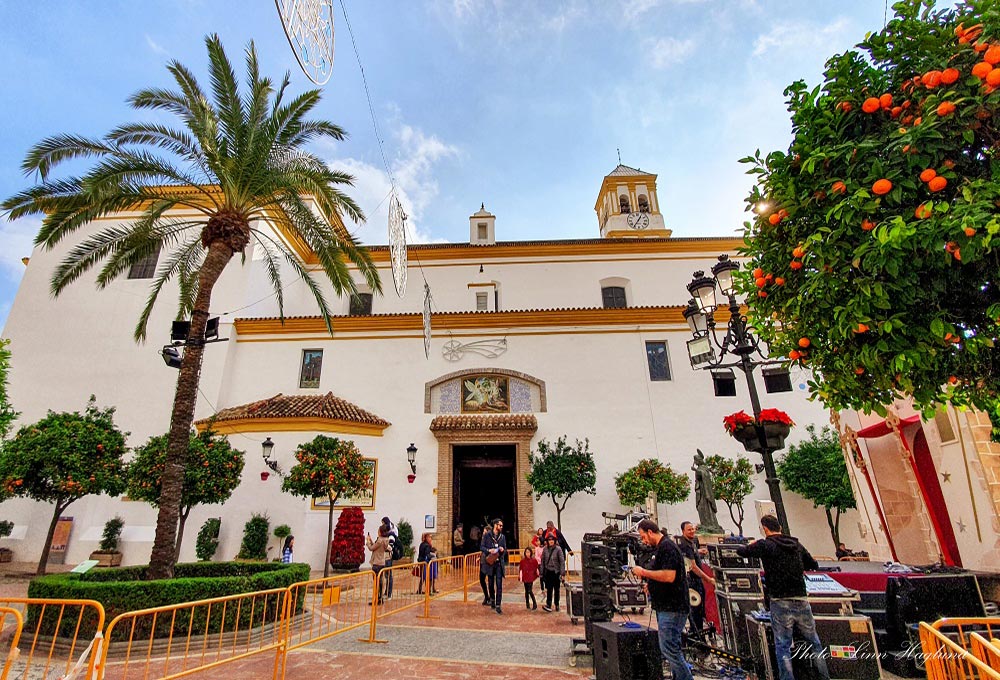 A white, historic church building with a palm tree, orange trees, several people including children and a stage with large sound system