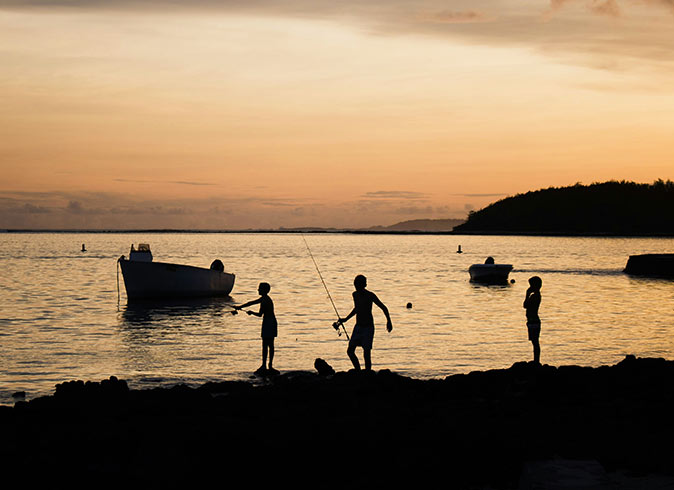 Life in Mauritius silhouettes of 3 kids fishing as the sun sets, with boats in the background