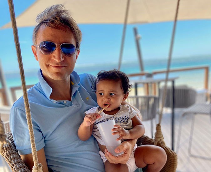 Philip with one of his children enjoying life in Mauritius A blond man with sunglasses, smiling, holding a small child, with a tropical beach in the background