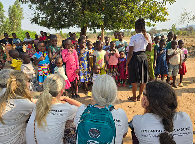 Visiting a local school while volunteering in Malawi 4 white women looking at a large group of African kids singing with a teacher in front of them