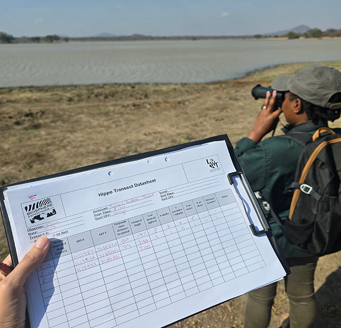 A hand holding a data sheet with a woman in the background looking at a lake through binoculars