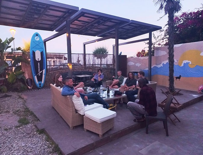 several people sitting on comfortable couches around a table in a backyard with a surfboard in the background