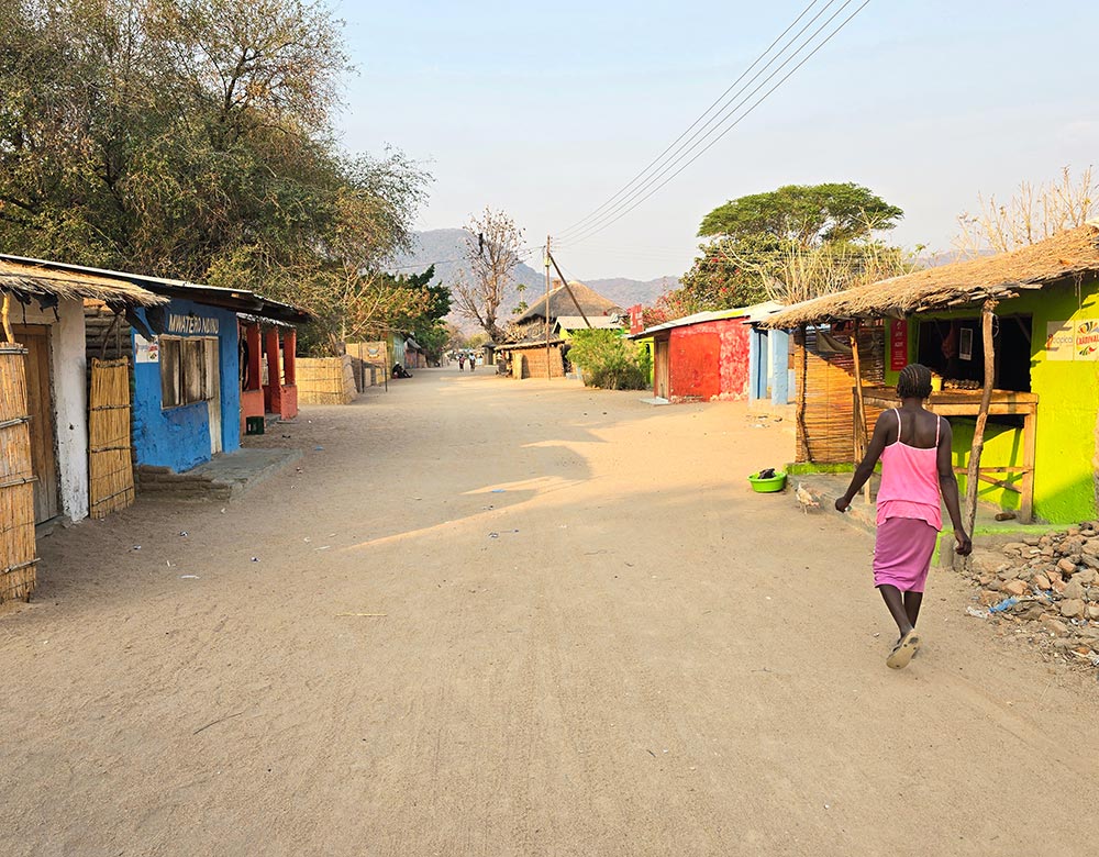 a woman in pink clothes walking on a sandy road with basic houses on both sides