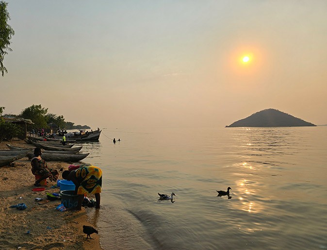 Cape Maclear on Lake Malawi sunset over a large lake with several ducks, a sandy beach with sever people and wooden canoes
