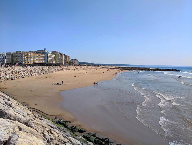 A coastal area liked by digital nomads in Portugal A long sandy beach with some people, a stone wall and a few highrise buildings in the background