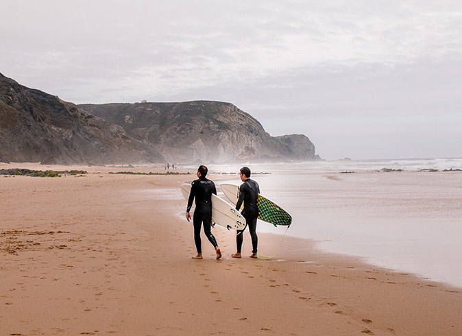 Surfers working remotely from Portugal 2 surfers walking on a sandy beach with their surfboards under their arms and some mountains in the background
