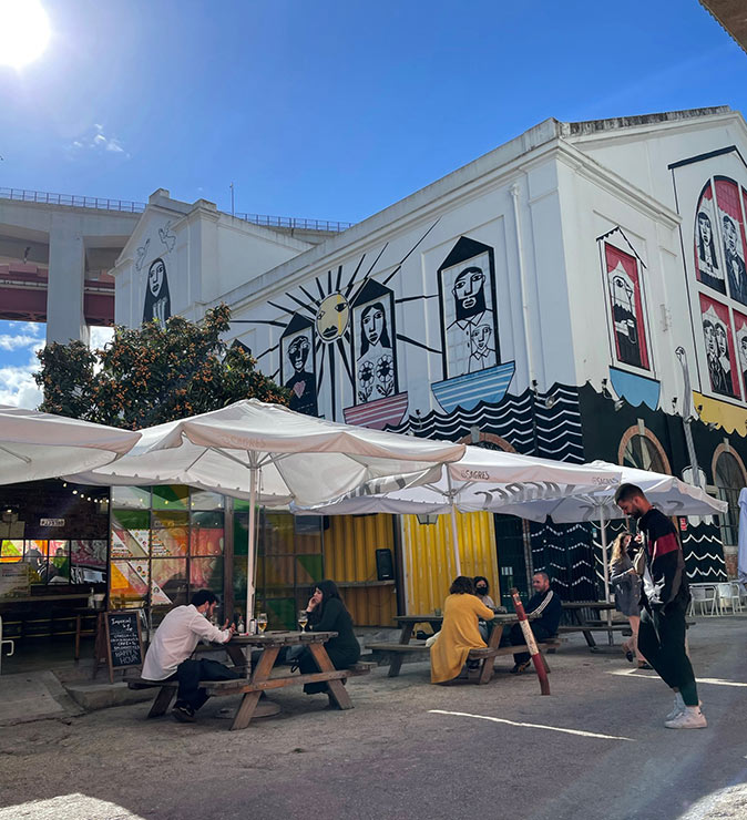 One of the best neighborhoods to live in Lisbon A city neighborhood with several picnic tables with umbrellas and several people, surrounded by white buildings with streetart
