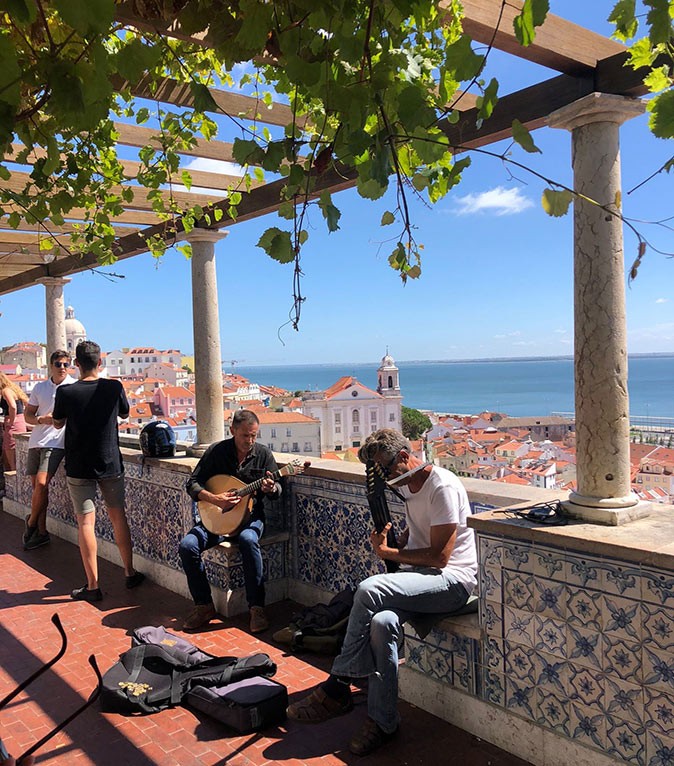 Where in Lisbon to live 2 men playing the guitar on a terrace with houses and the sea in the background