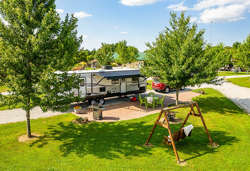 A large RV parked at an RV park surrounded by trees, a seating area and a bbq