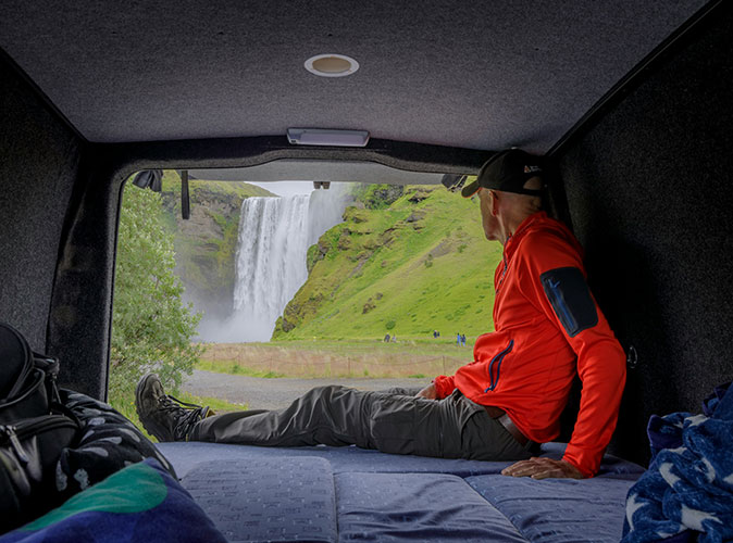 Exploring the beauty of Iceland in a camper A man sitting inside of an RV looking out towards a large waterfall surrounded by greenery