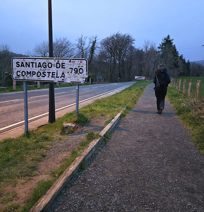 Walking the Camino de Santiago as a digital nomad A woman with a black backpack walking past a sign that reads 'Santiago de Compostela'