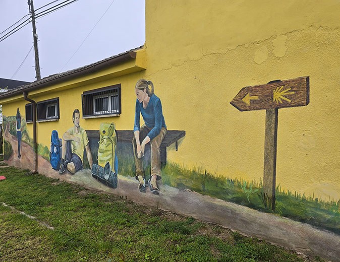 Street art along the Camino de Santiago A building painted yellow with street art showing several pilgrims, backpacks and a Camino sign