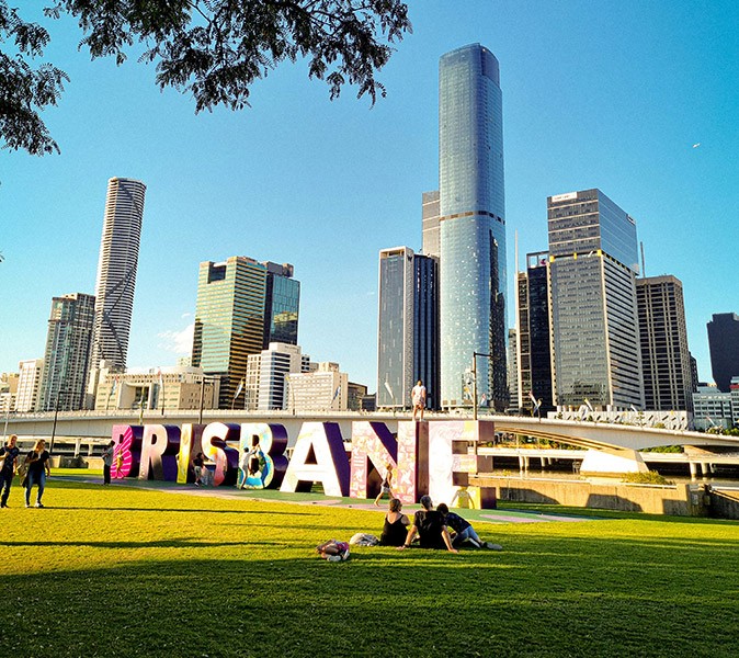 A green field with several people sitting in it, with large letters spelling 'Brisbane' and highrise in the background