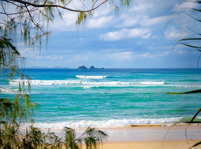 A sandy beach, gentle waves, bright blue water and several swimmers and surfers in the distance with islands showing in the background