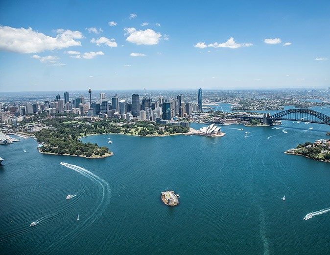 Aerial view of a city with highrise, green trees, water with several boats and a large bridge