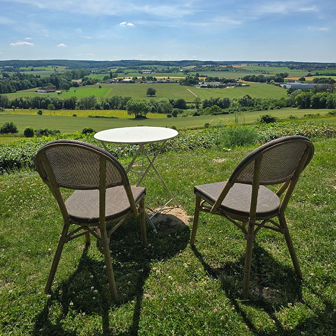 A small table with 2 chairs, overlooking a green valley with some buildings in the background