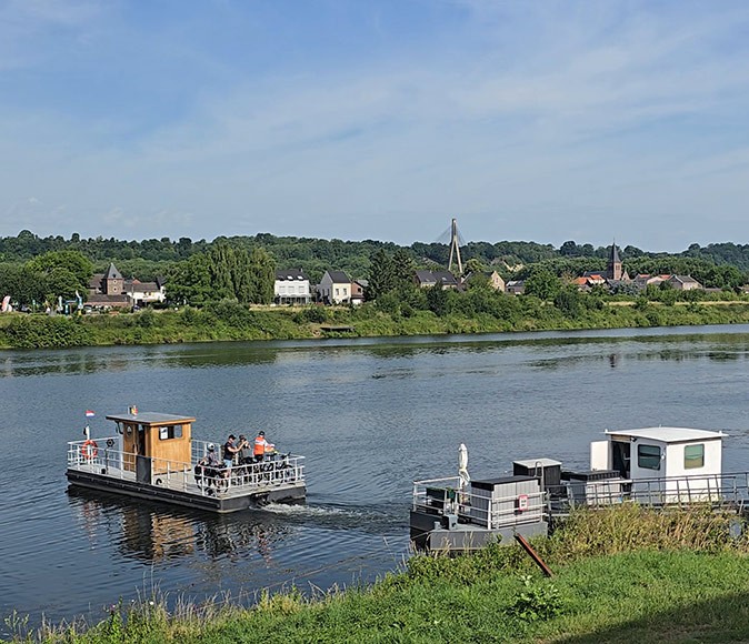 A ferry on a calm river with greenery in the foreground and greenery with some buildings in the background