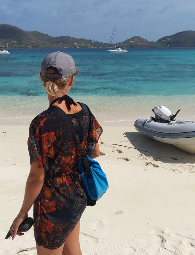 Sailing life in Grenada A woman in a dark dress and gray cap looking towards anchored boats while standing on a sandy beach with a dinghy