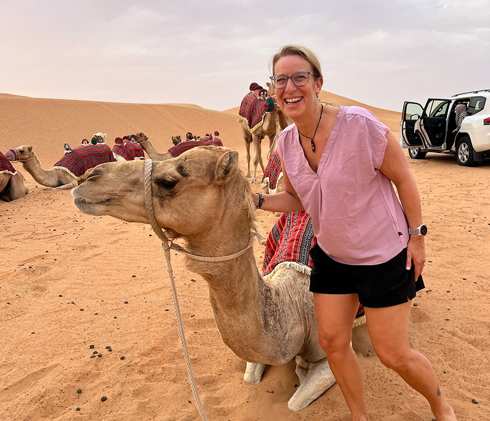 A blond woman over 50 standing next to a camel in a desert