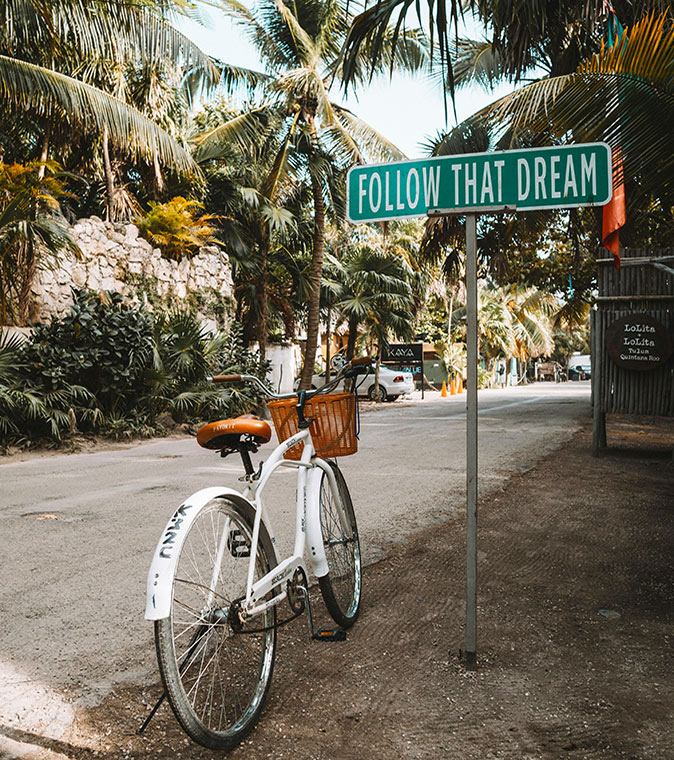 Follow that dream, travel at any age A green street sign with the text "Follow That Dream", a white bicycle next to it and tropical trees in the background