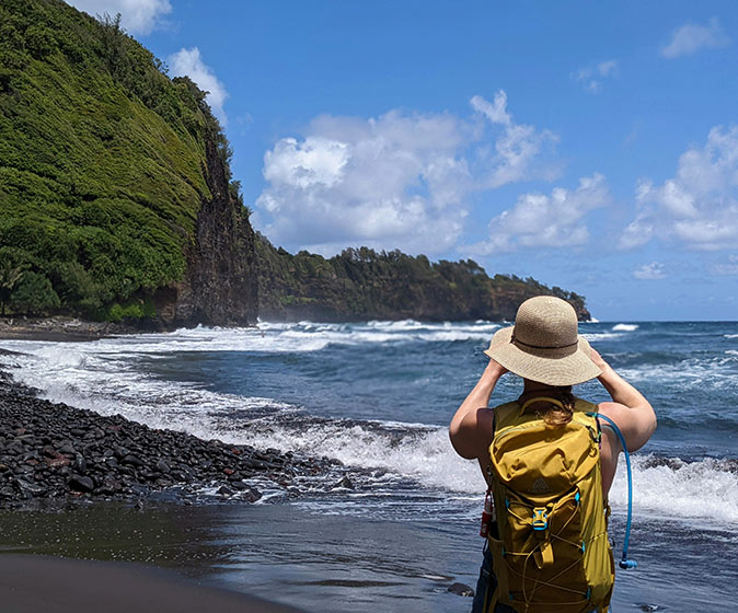 Solo travel on a budget for women over 50 A woman with a beige sun hat standing on a beach looking at the waves with a high cliff covered in greenery in the background