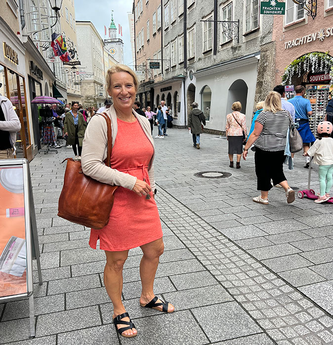 Exploring Europe solo as a woman over 50 A blond woman smiling at the camera in a typical European city with a gray paved roads, several pedestrians and historic buildings