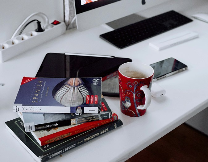 Learning Spanish while living in Spain A pile of Spanish language learning books on a white desk with a coffee cup and a cumputer