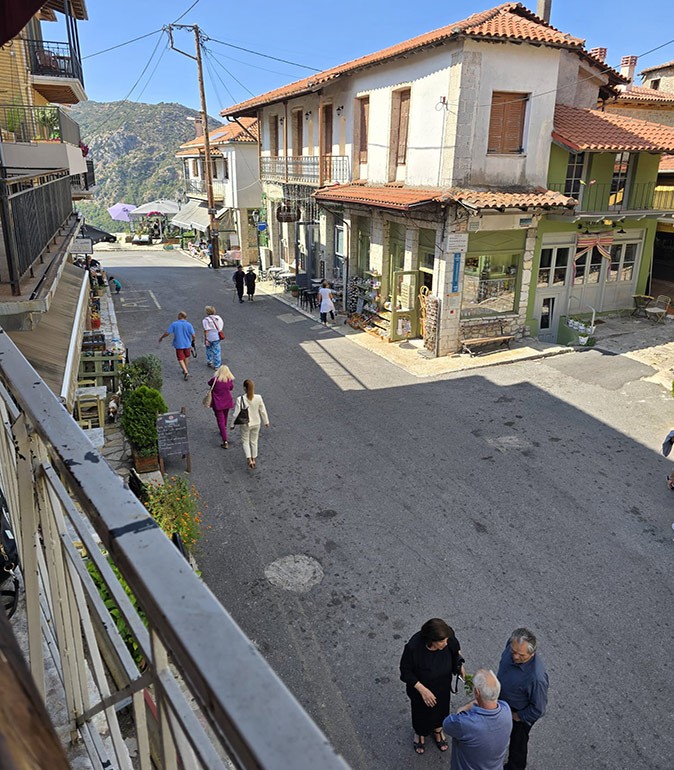 A town in the Peloponnese region of Greece View of a small street in a historic Greek town showing several people walking, gray and brown buildings with some shops and restaurants