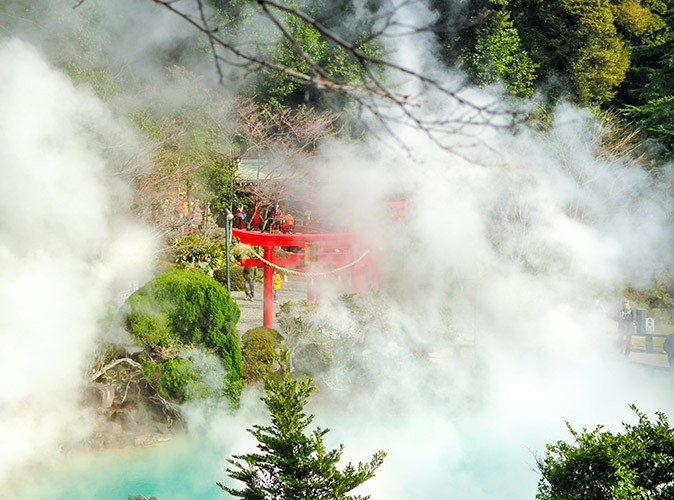 One of the best onsen towns to visit in Japan Steam rising from hotsprings with a red shrine and greenery in the background