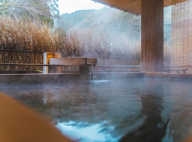 One of many onsen in Japan A square bath, outdoors, with steam rising up from it and part of a mountain and some bushes in the background