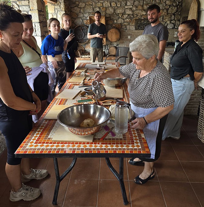 Cultural immersion and community building while working remotely from Greece An older Greek woman teaching an international group of people how to make Greek bread, stainding around a table in a stone building