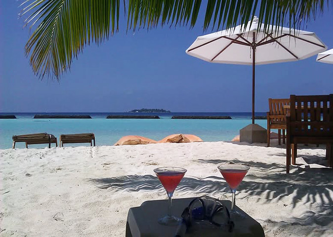 2 cocktails on a small white table with a snorkeling mask, whitel sandy beach and bright blue ocean in the background