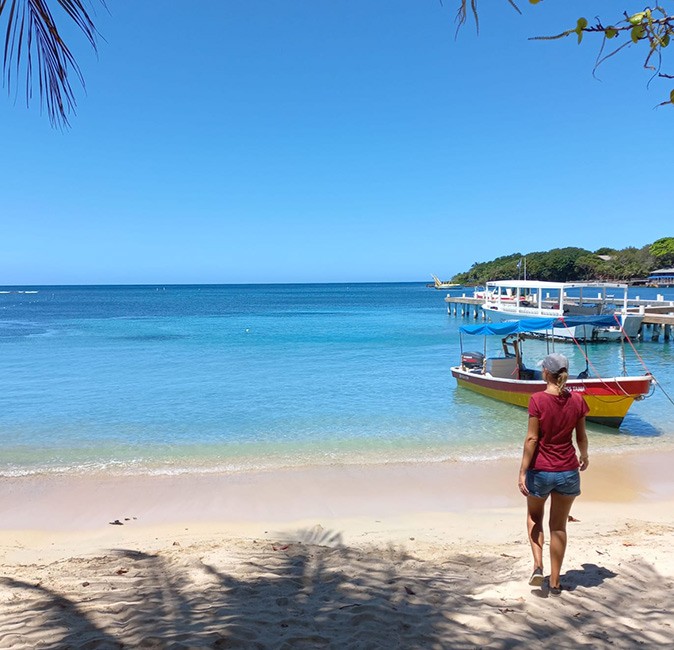 A woman on a white sandy beach walking towards the water with a colorful wooden boat and part of a palm tree showing