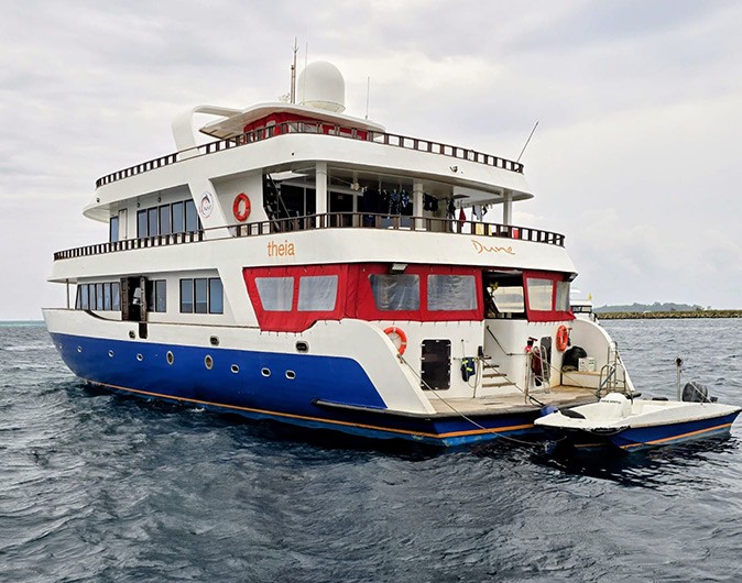 A liveaboard at sea in the maldives with a white and blue hull, some red window covers and a small motorboat tied behind it
