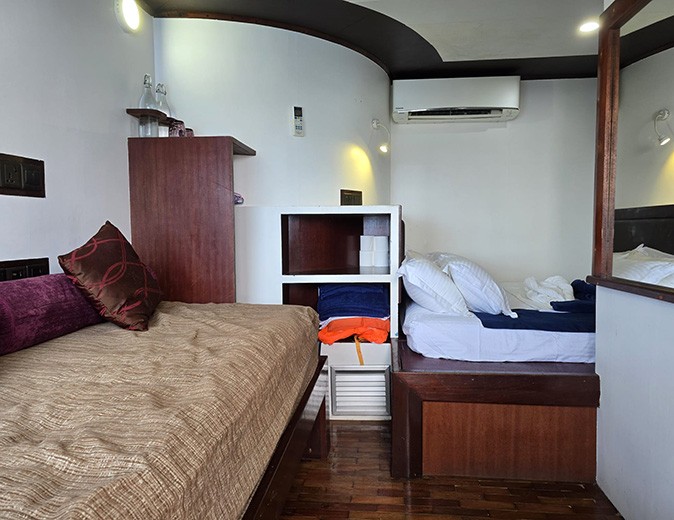 A cabin on a Maldivian liveaboard showing a brown sofa, a white double bed, some shelves and some cushions
