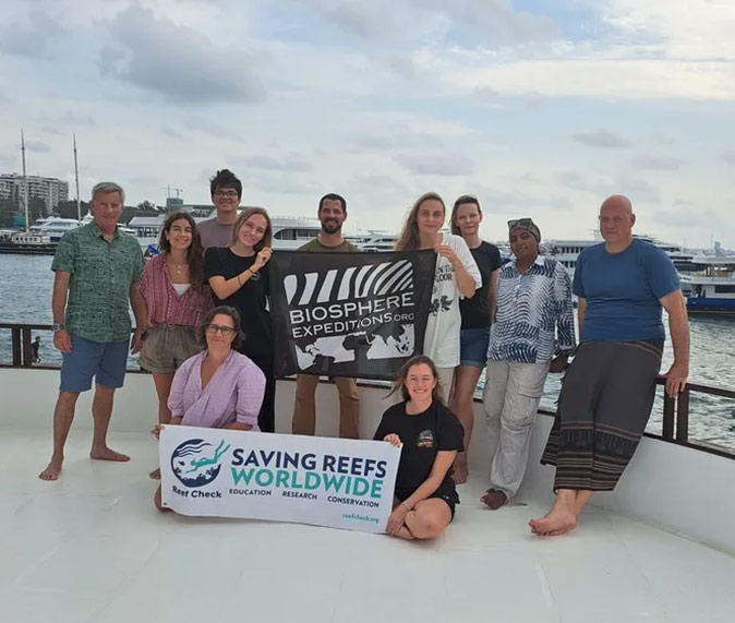 A group picture on a liveaboard boat in the Maldives with 2 women holding a banner that reads 'saving reefs worldwide' and another banner that reads 'Biosphere expeditions'