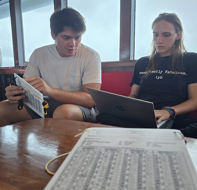 A man and a woman sitting on a red couch inside a boat looking at a laptop screen while the man holds a data sheet