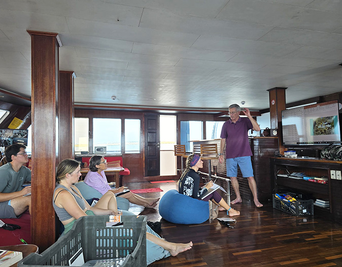 A group of people sitting inside a boat looking at a man giving instructions next to a tv screen