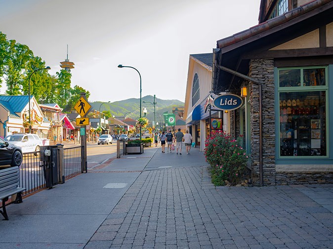 A guide for visiting Gatlinburg An urban setting with a gray sidewalk, a road, and parts of shops, with several people walking on the sidewalk
